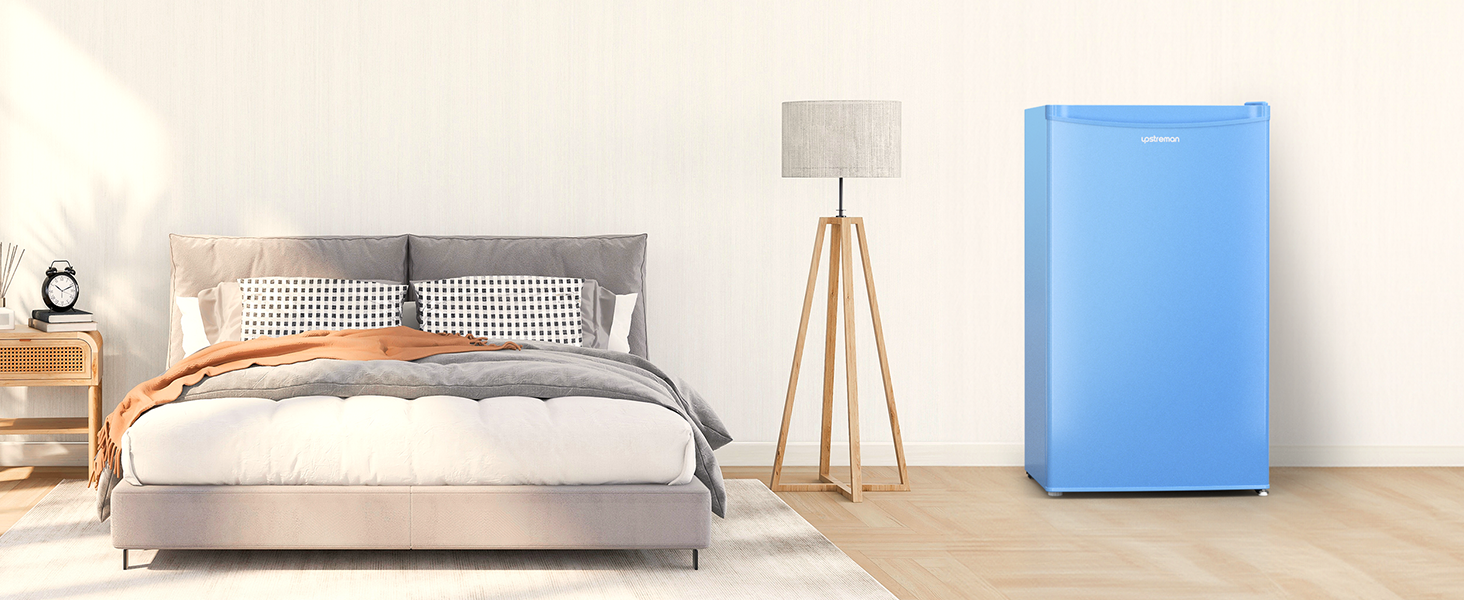 Intérieur de la chambre avec lit rembourré gris, table de chevet en bois, lampadaire trépied et réfrigérateur compact bleu clair. Des planchers de bois franc et des murs blancs complètent le décor.